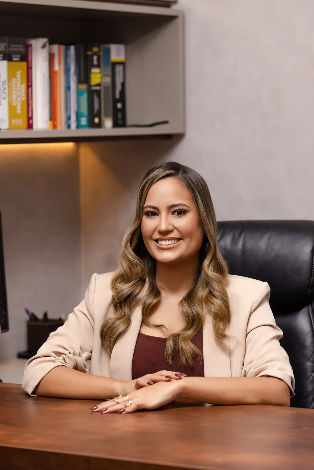 Emylly Belmiro sentada à mesa em seu escritório, com estante de livros jurídicos ao fundo